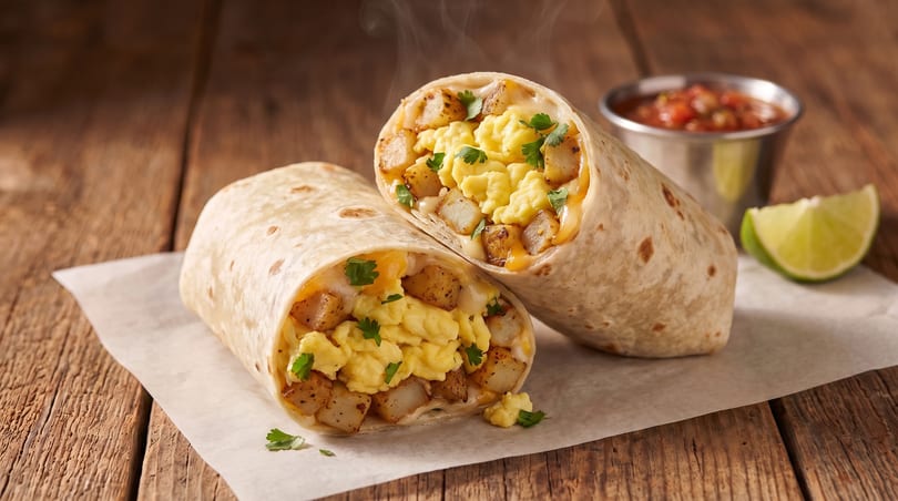 Two steaming breakfast burritos filled with scrambled eggs, potatoes, cheese, and parsley, with salsa and lime.